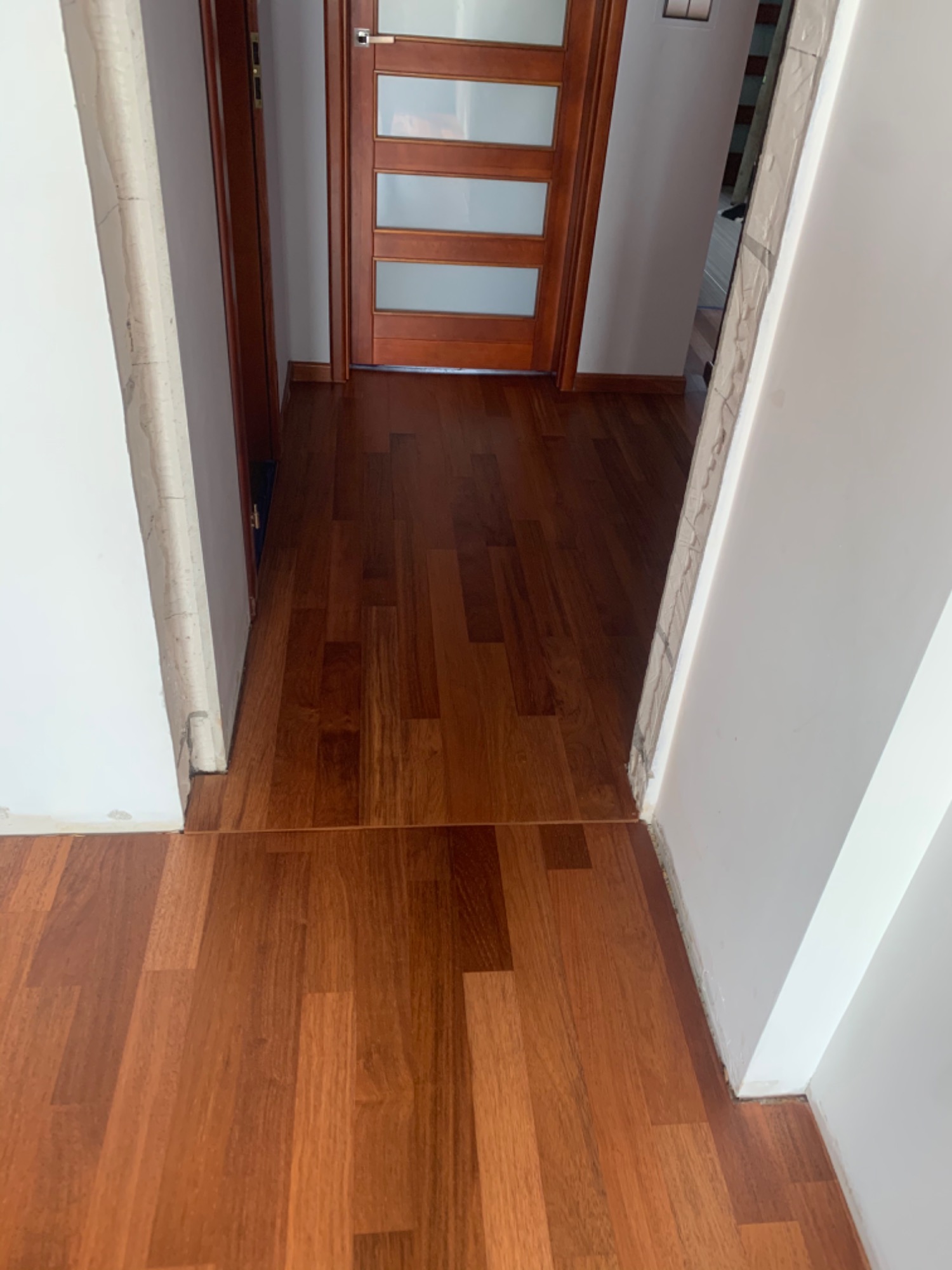 Nowo ułożona, brązowa podłoga z paneli drewnopodobnych w korytarzu, widoczne niedokończone wykończenia przy ścianach i ościeżnicy drzwiowej.