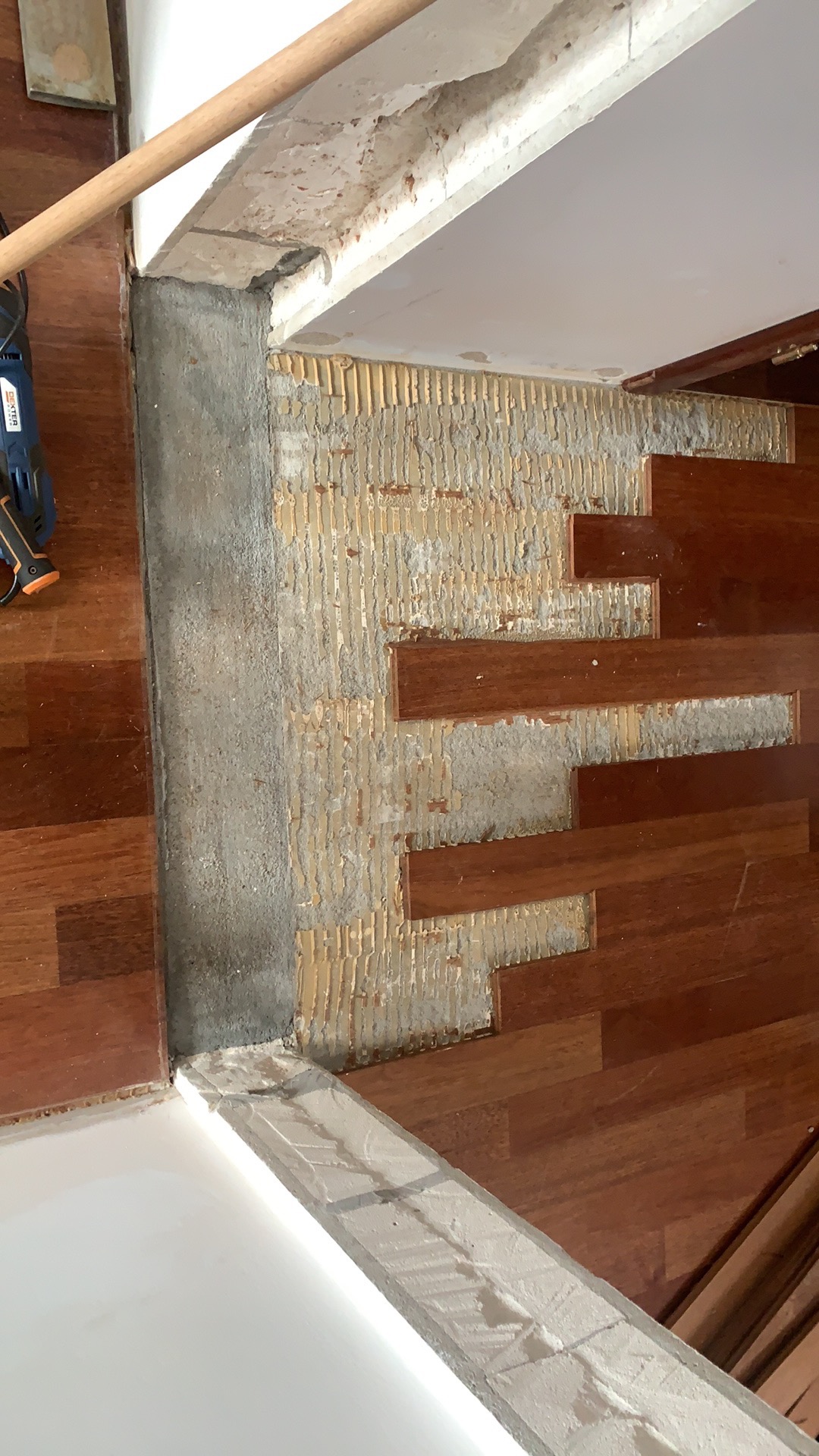 Fragment podłogi z ciemnego drewna, w trakcie prac wykończeniowych, widoczne niedociągnięcia przy ścianie i resztki kleju, obok leży niebieska wiertarka.