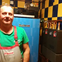 Uśmiechnięty instalator w roboczym ubraniu na tle niebieskiego pieca Buderus i miedzianych rur w pomieszczeniu z żółto-niebieskimi kafelkami.