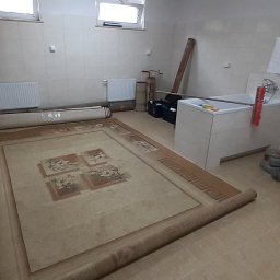 Aqua AS - Duży, zwinięty dywan z wzorem kwiatowym leży na podłodze w pomieszczeniu z białą umywalką, grzejnikami i sprzętem do czyszczenia, w tle zwinięty drugi dywan zabezpieczony folią.