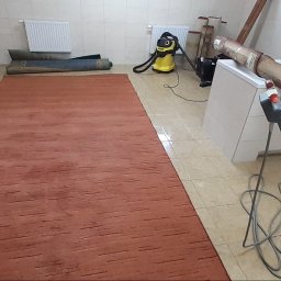 Aqua AS - Pomarańczowy dywan z krótkim włosiem rozłożony na kafelkach w łazience, obok zwinięte dywany, odkurzacz i maszyna do czyszczenia dywanów z widocznym kablem.