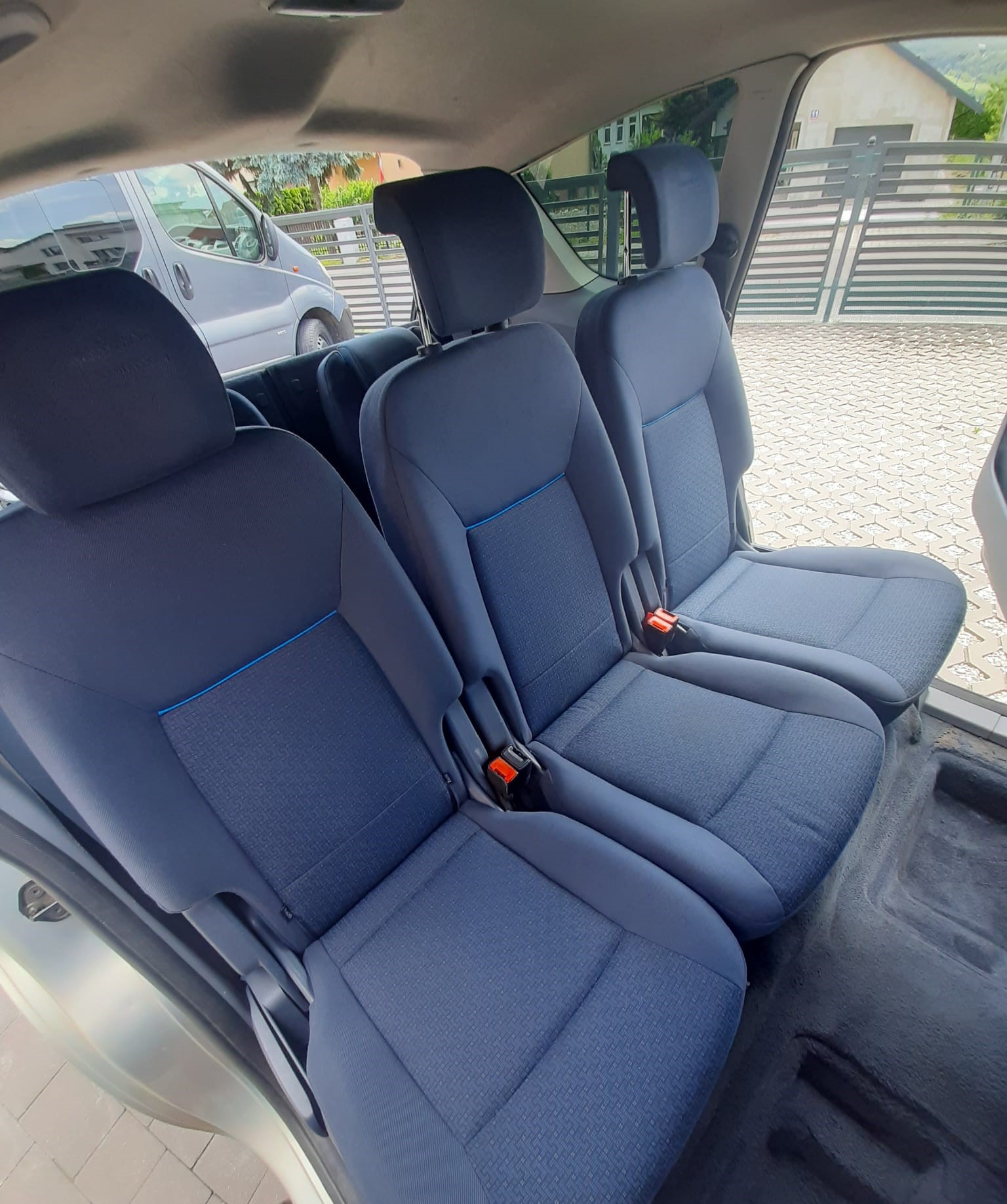 Wnętrze samochodu z trzema rzędami czystych, niebieskich foteli z pasami bezpieczeństwa, widok z perspektywy drzwi, z zewnątrz widoczny fragment chodnika i budynku.