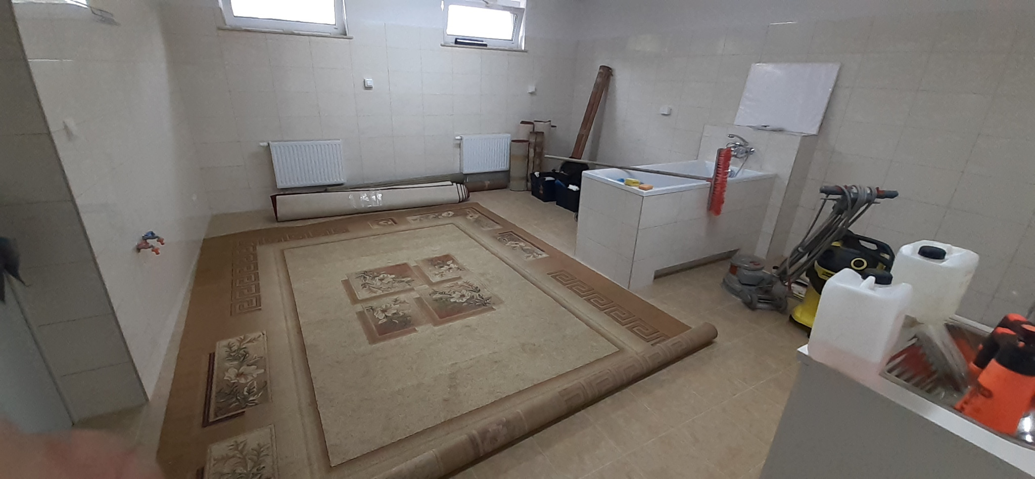 Duży, zwinięty dywan z wzorem kwiatowym leży na podłodze w pomieszczeniu z białą umywalką, grzejnikami i sprzętem do czyszczenia, w tle zwinięty drugi dywan zabezpieczony folią.