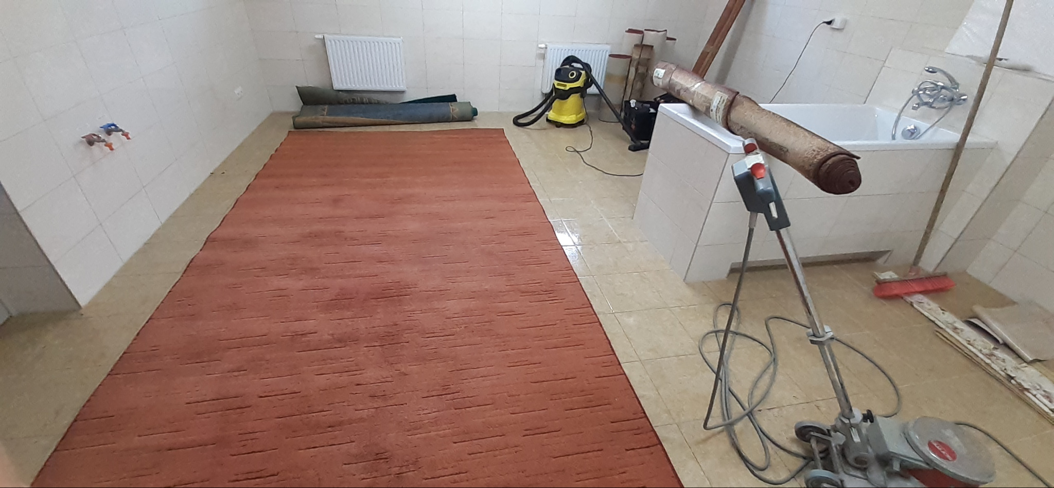 Pomarańczowy dywan z krótkim włosiem rozłożony na kafelkach w łazience, obok zwinięte dywany, odkurzacz i maszyna do czyszczenia dywanów z widocznym kablem.