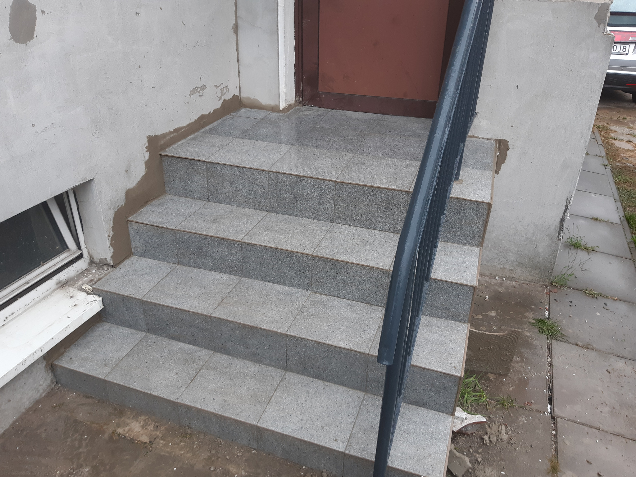 Wykończone schody wejściowe do budynku z szarych płytek, z metalową poręczą po prawej stronie, ściany budynku w trakcie naprawy, widoczne ślady po łataniu.