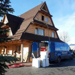 Drewniany dom w stylu góralskim z niebieskim samochodem dostawczym firmy izolacyjnej, z którego przez okno poddasza wystaje wąż do wdmuchiwania materiału izolacyjnego. Na zewnątrz, obok samochodu...
