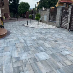 Ułożona, szaro-niebieska kostka brukowa na podjeździe, z okrągłym klombem z białych kamieni i dwoma małymi drzewkami, brama z drewnianymi elementami w tle.