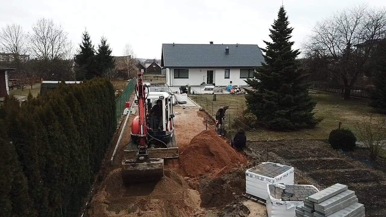 Prace ziemne pod budowę chodnika z kostki brukowej: Mini koparka wykopuje rów, obok stos kostki brukowej na palecie, w tle dom jednorodzinny i pracownicy z łopatami.