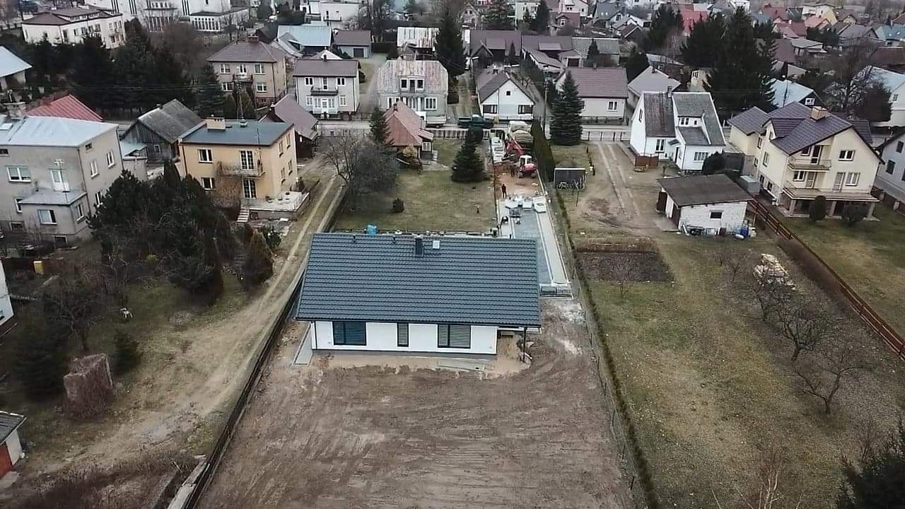 Widok z drona na nowo wybudowany dom z szarą dachówką i białymi ścianami, w trakcie prac związanych z zagospodarowaniem terenu, widoczna ułożona ścieżka z kostki brukowej i przygotowany teren...