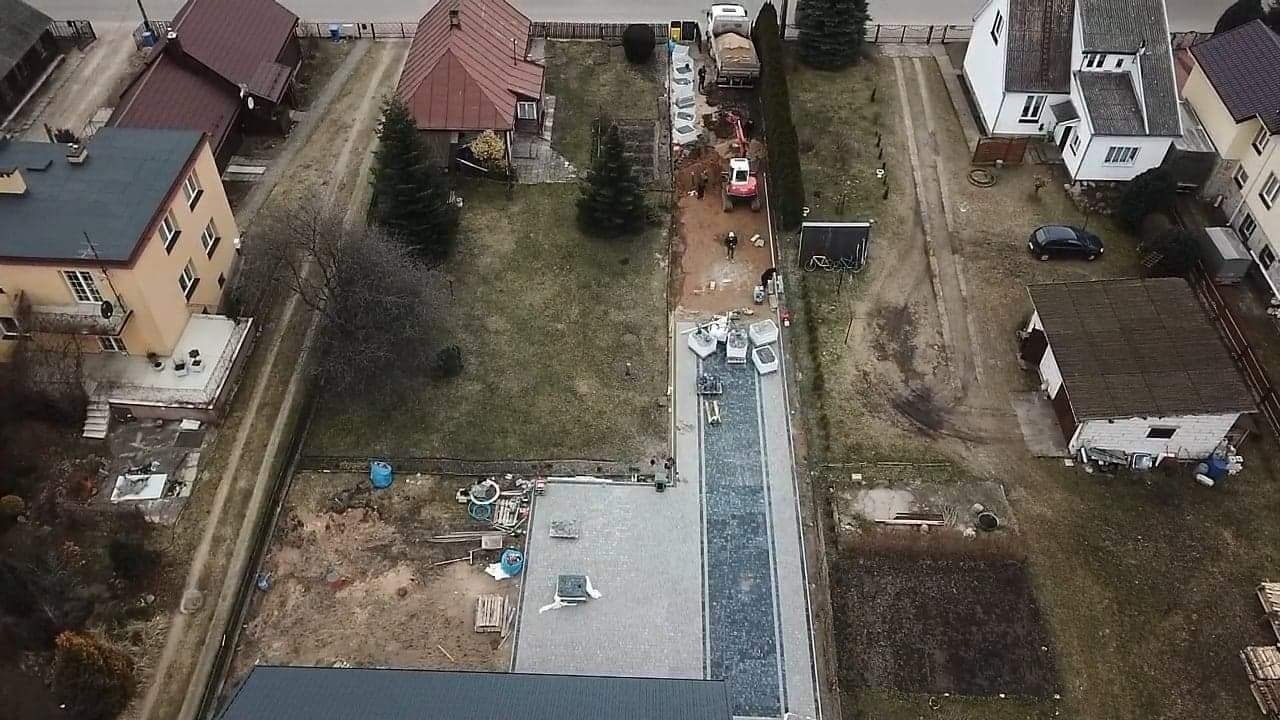 Widok z góry na budowę podjazdu z kostki brukowej, częściowo ułożonej w szaro-niebieskie wzory, z widoczną koparką i materiałami budowlanymi na działce.