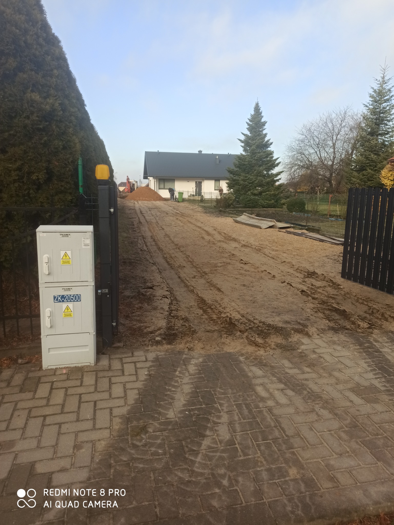 Teren przed domem w trakcie przygotowywania podłoża, widoczne ślady opon, hałda piasku i maszyna budowlana w tle, przy ogrodzeniu skrzynka elektryczna z ostrzeżeniami.