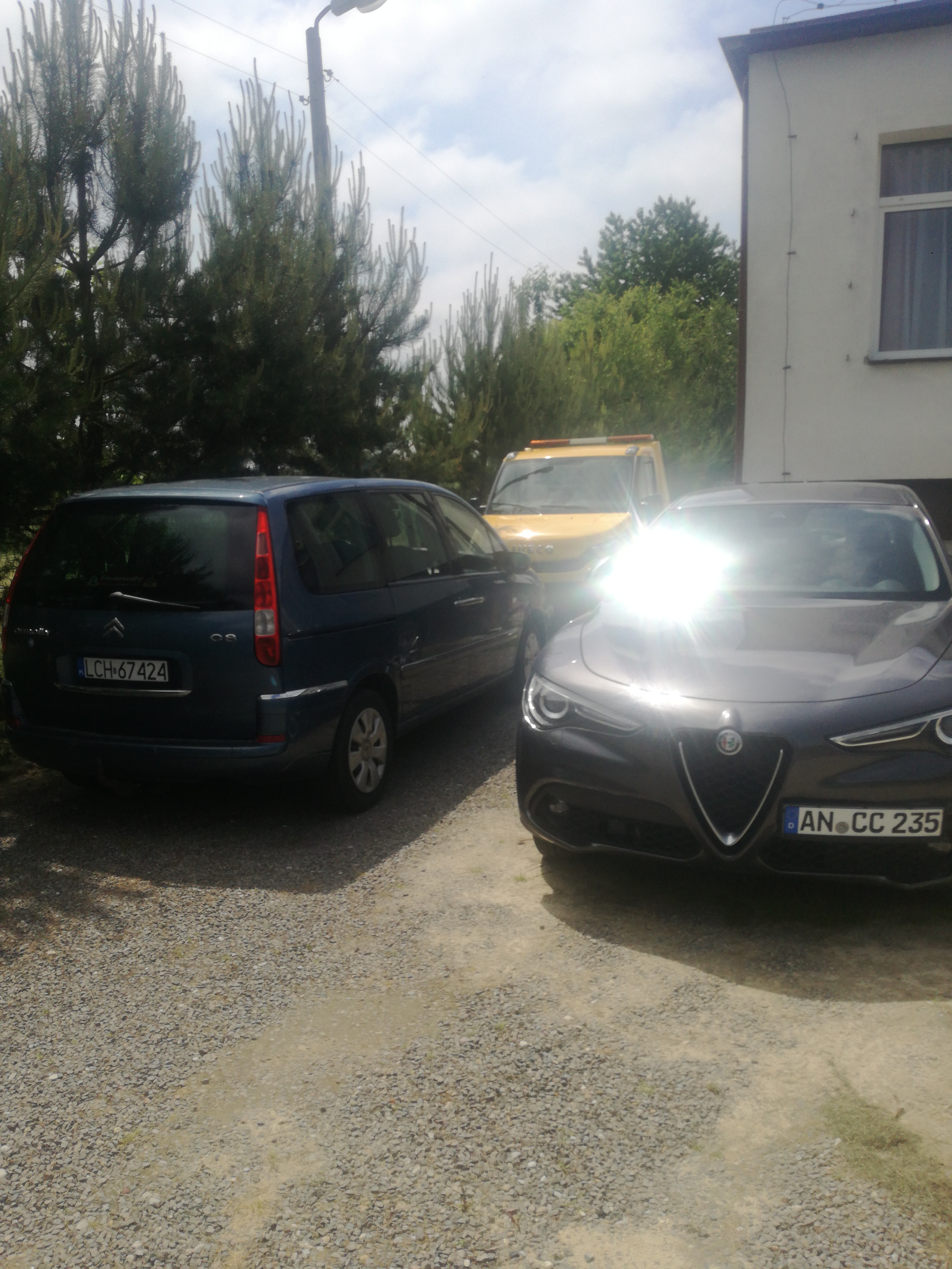 Alfa Romeo i Citroen stoją obok siebie na żwirowym podłożu, w tle widoczna żółta laweta i budynek z białego tynku.