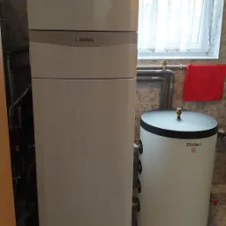 Biała pompa ciepła Vaillant z zasobnikiem ciepłej wody w kotłowni, widoczne rury i okno z firanką.