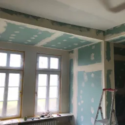 Pokój w trakcie remontu: ściany i sufit pokryte płytami gipsowo-kartonowymi z widocznymi miejscami szpachlowania, okna zabezpieczone folią malarską, drabina rozstawiona przy ścianie, narzędzia...