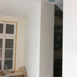 Bartosz BB - Wnętrze w trakcie prac wykończeniowych: okno zabezpieczone folią, ściany częściowo pomalowane na biało, widoczne ślady po szpachlowaniu na suficie, butelka wody i narzędzia na parapecie.