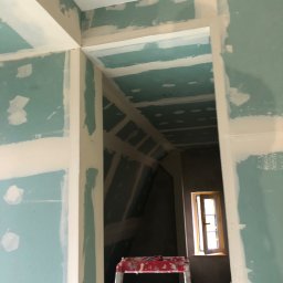 Bartosz BB - Wnętrze w trakcie prac wykończeniowych: ściany i sufit pokryte płytami gipsowo-kartonowymi, widoczne łączenia wymagające szpachlowania, drabina malarska w tle, okno z drewnianą ramą w głębi...