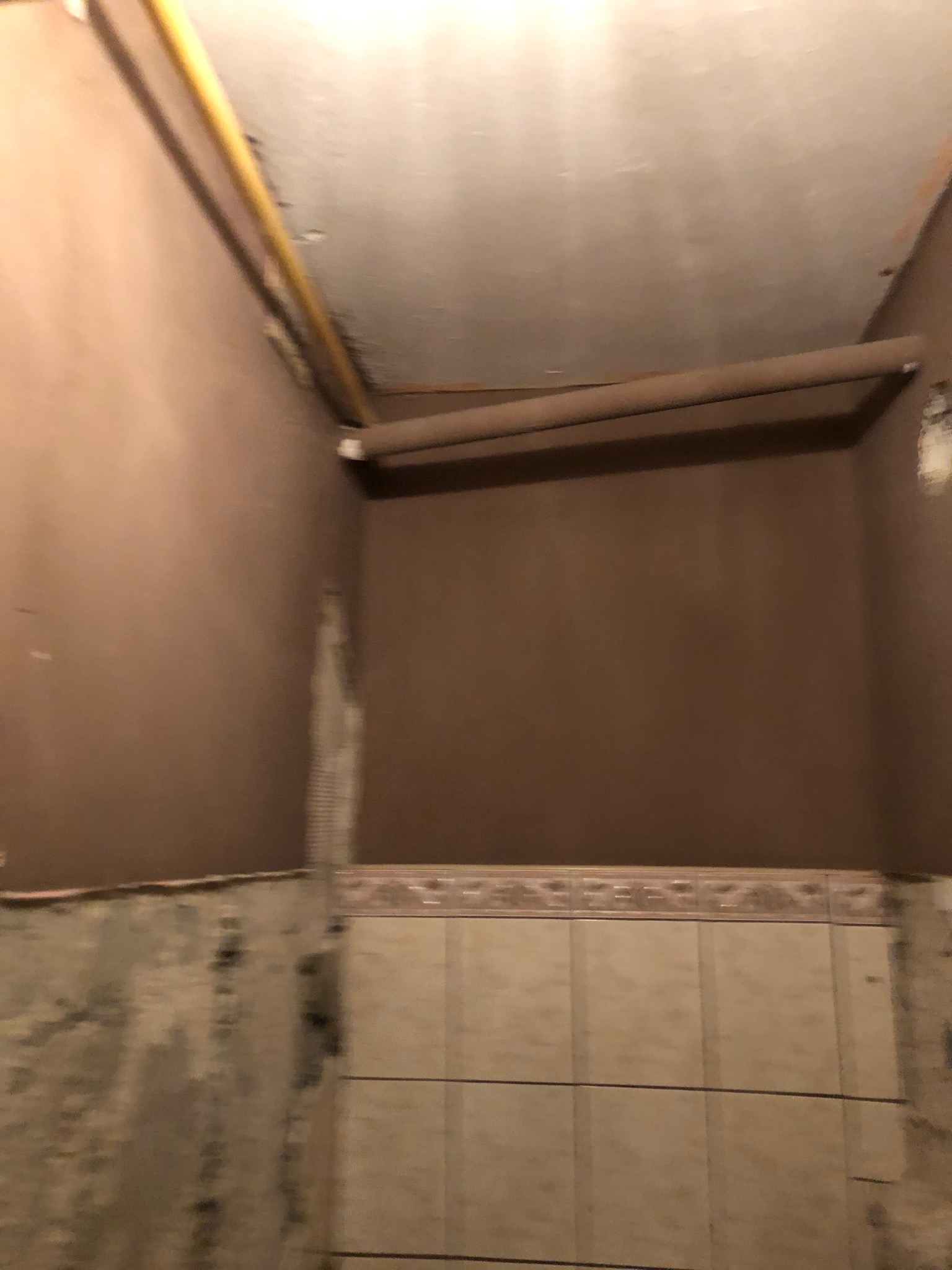 Wnętrze w trakcie prac wykończeniowych: częściowo położone płytki, świeżo pomalowane ściany w odcieniach brązu, widoczne rury pod sufitem i fragmenty niezabezpieczonej ściany.