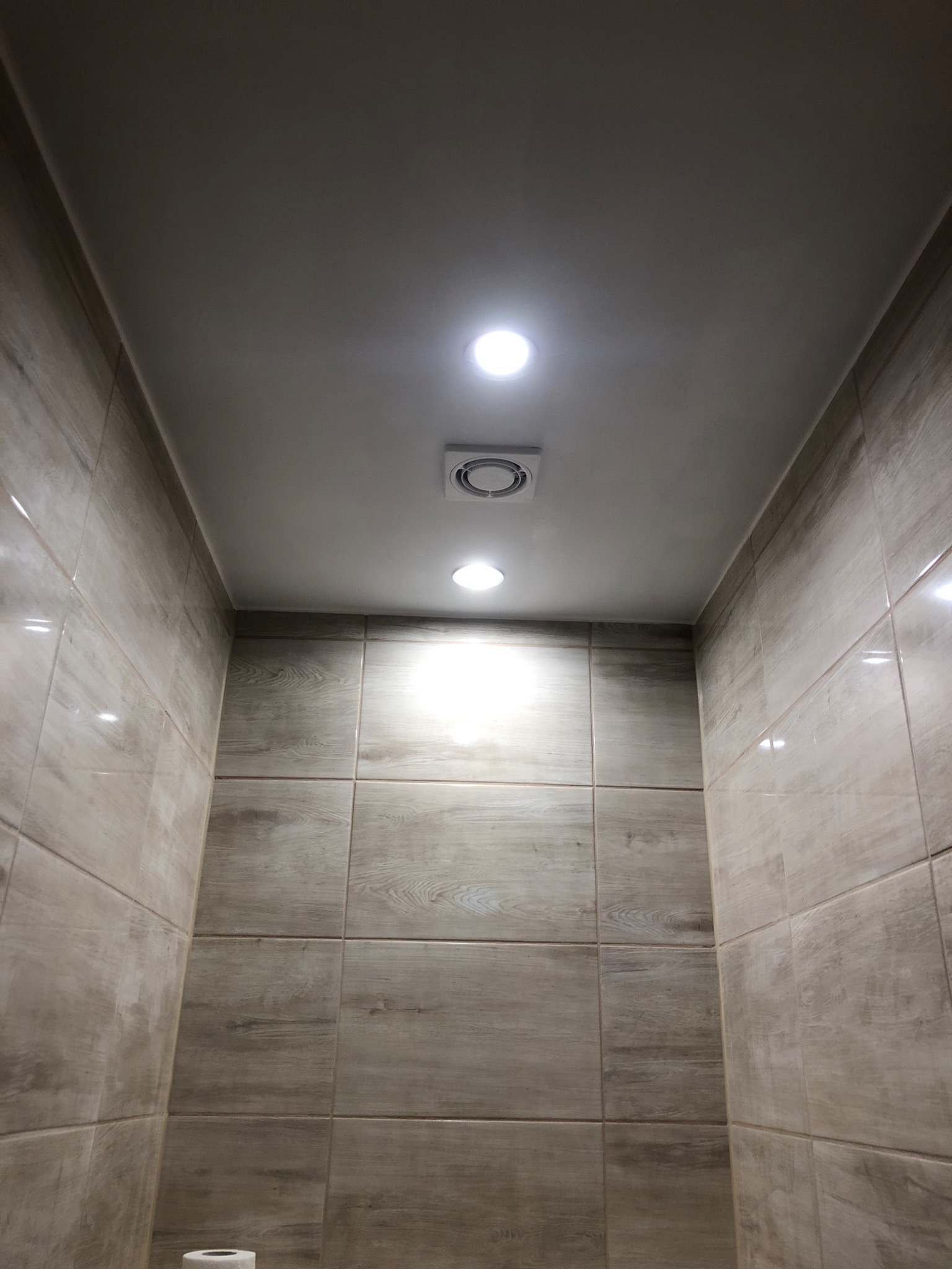 Wnętrze wykończonej łazienki z beżowymi płytkami imitującymi drewno, oświetleniem punktowym w suficie i kratką wentylacyjną.