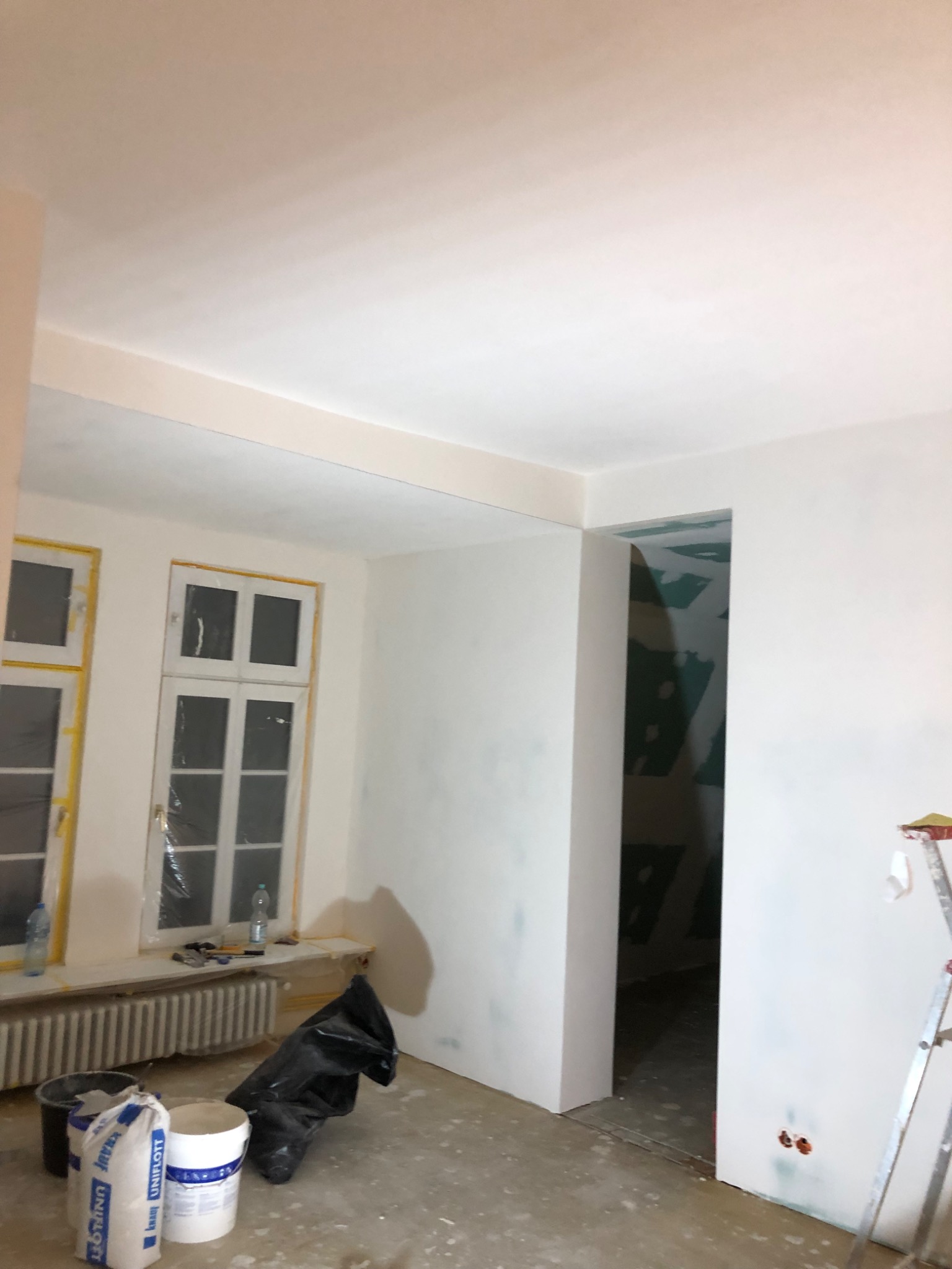 Pokój w trakcie prac wykończeniowych, ściany świeżo pokryte gładzią, okna zabezpieczone folią, widoczne worki z zaprawą, wiadro i drabina.