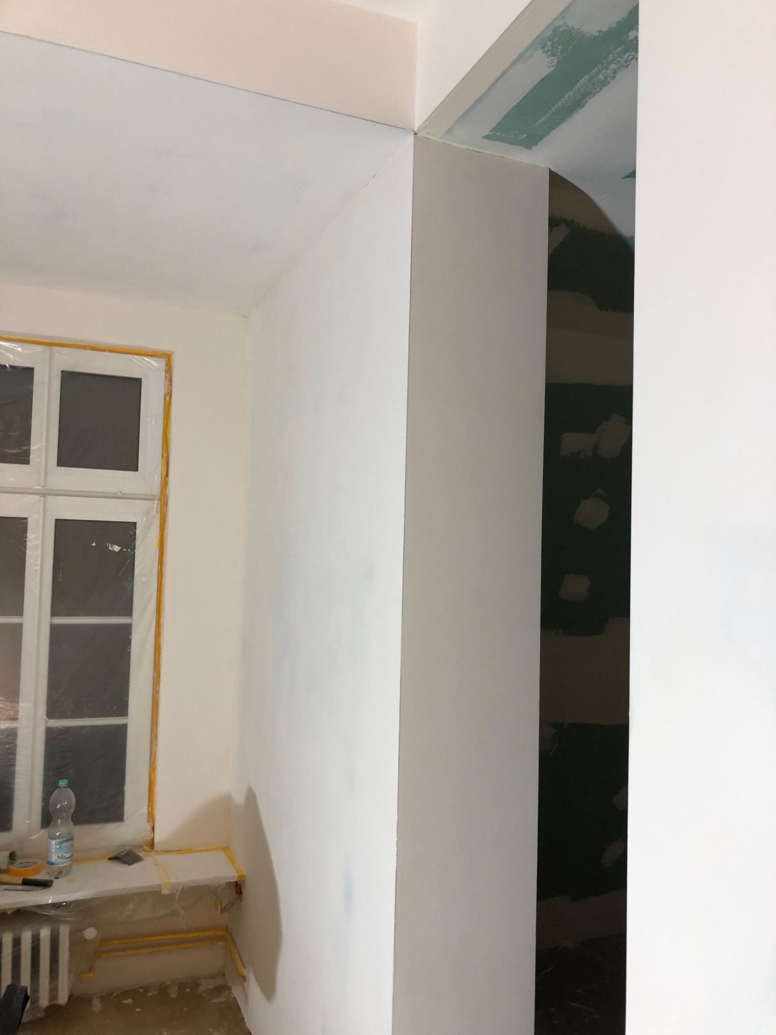 Wnętrze w trakcie prac wykończeniowych: okno zabezpieczone folią, ściany częściowo pomalowane na biało, widoczne ślady po szpachlowaniu na suficie, butelka wody i narzędzia na parapecie.
