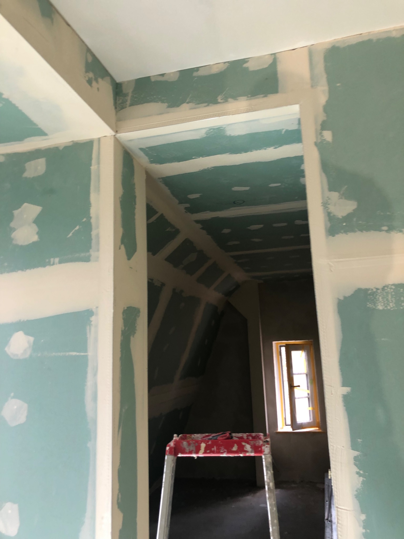 Wnętrze w trakcie prac wykończeniowych: ściany i sufit pokryte płytami gipsowo-kartonowymi, widoczne łączenia wymagające szpachlowania, drabina malarska w tle, okno z drewnianą ramą w głębi...