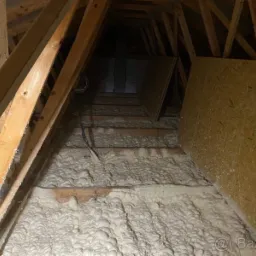 Poddasze z drewnianą konstrukcją pokryte warstwą jasnej pianki izolacyjnej, widoczne przewody elektryczne i płyta OSB.