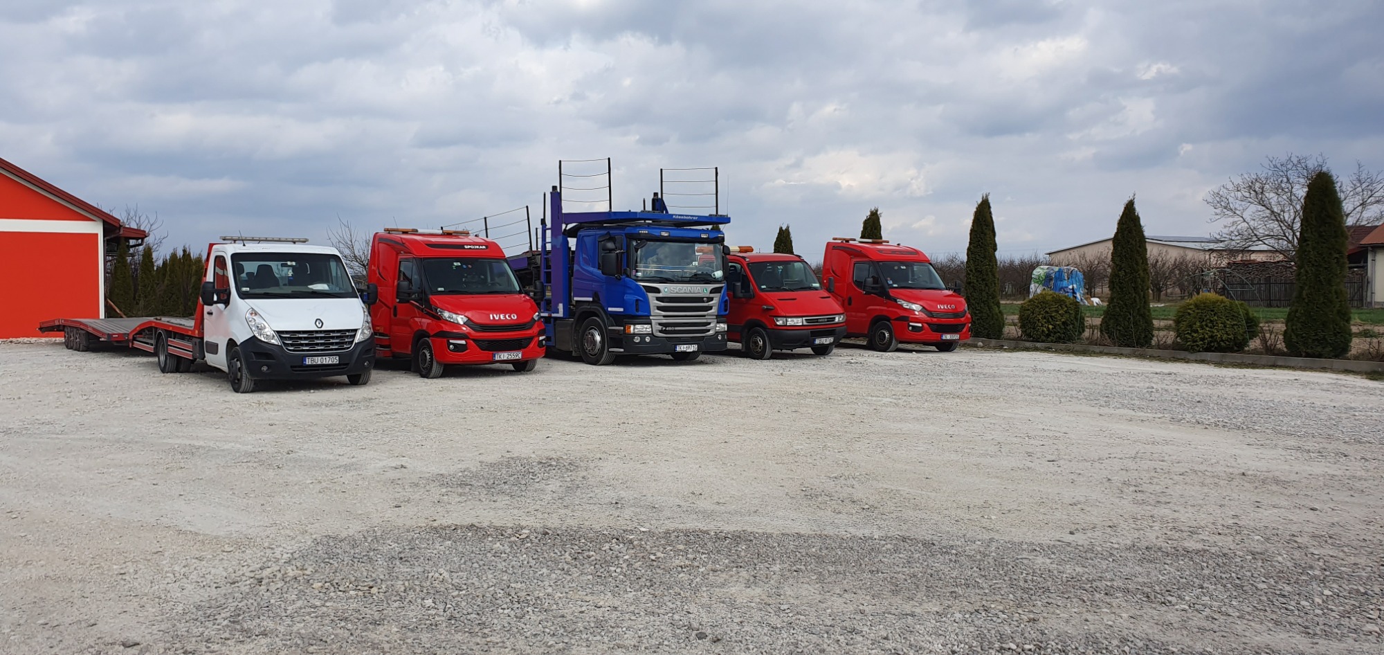 Flota lawet różnych marek (Renault, Iveco, Scania) w kolorach białym i czerwonym, zaparkowana na żwirowym placu pod pochmurnym niebem, z widocznym czerwonym budynkiem w tle.