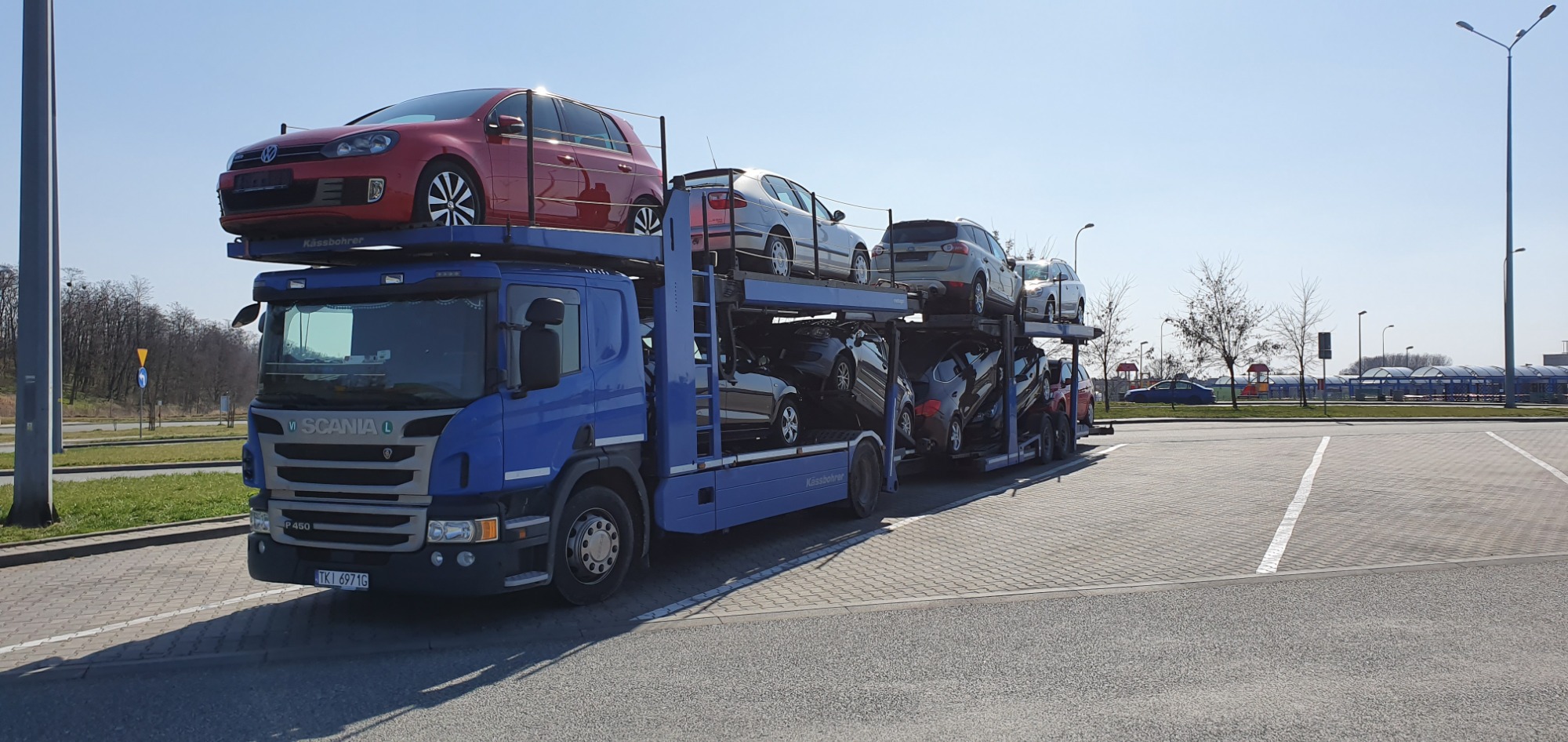 Niebieska laweta Scania P450 z załadowanymi samochodami osobowymi na parkingu, widok z przodu na tle słonecznego nieba i drzew bez liści.