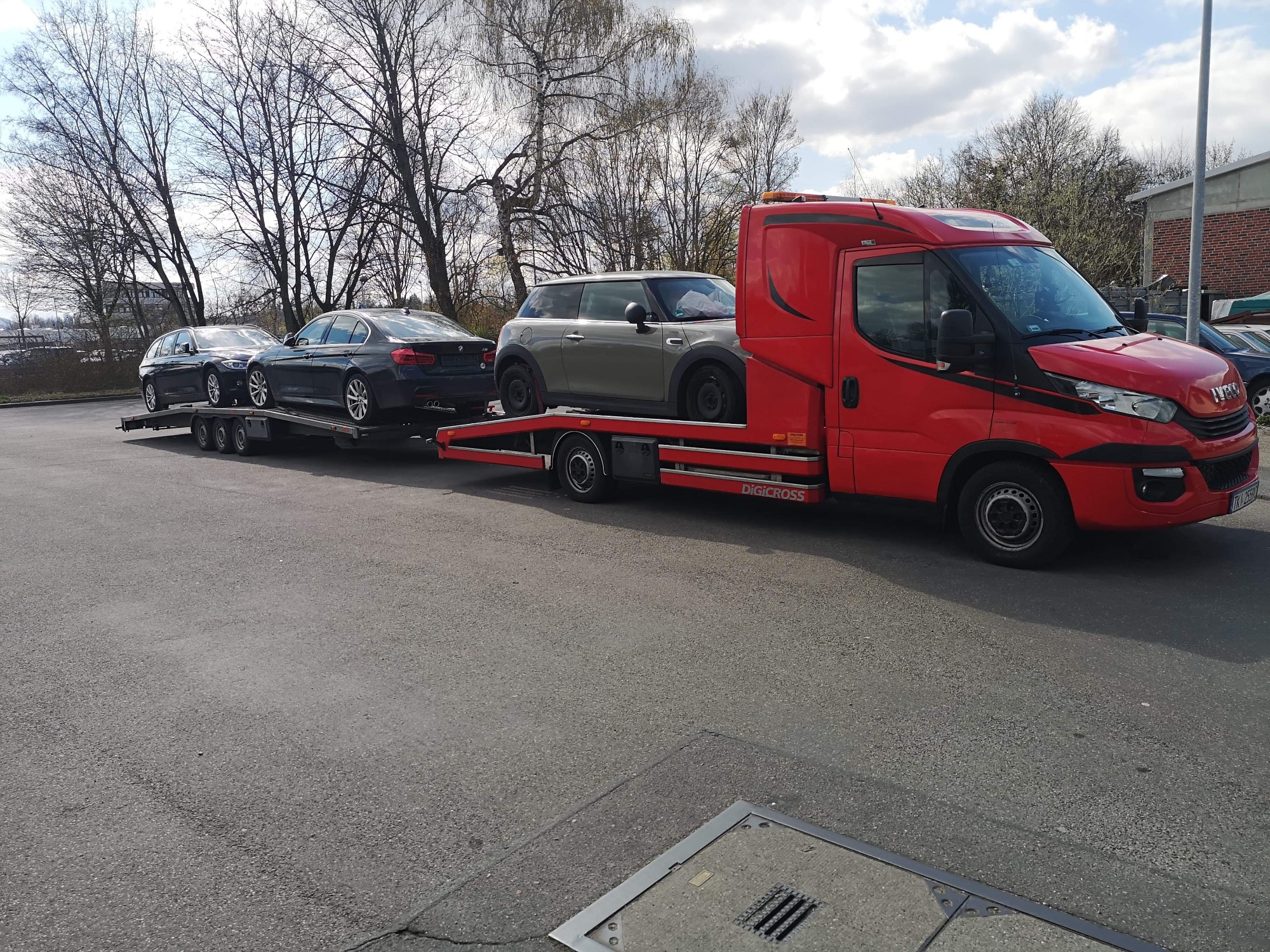 Czerwona laweta z trzema samochodami (dwa ciemne sedany i szary Mini Cooper) na platformie, zaparkowana na asfalcie w pochmurny dzień.