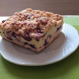 Anna jaskółka - Kwadratowy kawałek ciasta z kruszonką i owocami (prawdopodobnie borówkami) na białym talerzu, umieszczony na zielonej, teksturowanej podkładce.