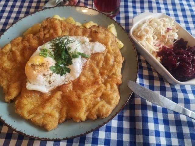 Sznycel wiedeński z jajkiem sadzonym i koperkiem na talerzu, obok surówka z kapusty i buraków na tle niebiesko-białej kraty.