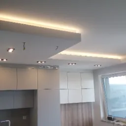 Nowoczesna kuchnia z białymi szafkami, drewnianymi akcentami i podwieszanym sufitem z oświetleniem LED, widoczne niedokończone podłączenie kabli.