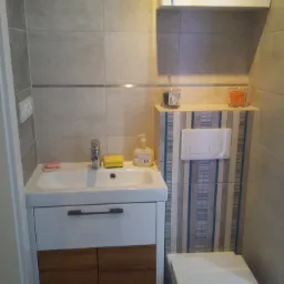 Nowoczesna łazienka z umywalką nablatową na szafce z drewnianymi frontami, wiszącą szafką, i toaletą, wykończona szarymi płytkami z dekoracyjnym pasem za toaletą.