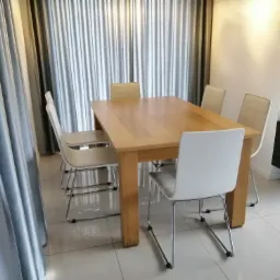 Jasny drewniany stół jadalny z krzesłami o metalowych nogach i białych siedziskach, ustawiony na błyszczącej, jasnej podłodze, z zasłonami w tle i rośliną w donicy po prawej stronie.