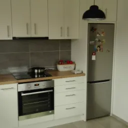 Jasna kuchnia z białymi szafkami, drewnianym blatem i szarą glazurą, z widocznym garnkiem na płycie indukcyjnej i dekoracjami na lodówce.