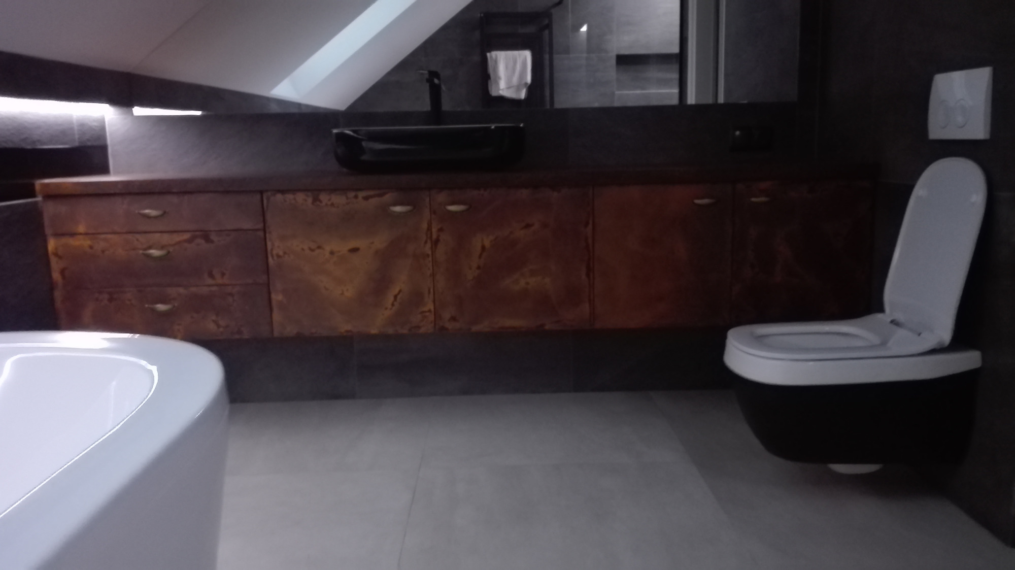 Nowoczesna łazienka w odcieniach szarości z drewnianą szafką pod umywalką, czarną umywalką nablatową i podwieszaną toaletą z otwartą klapą.