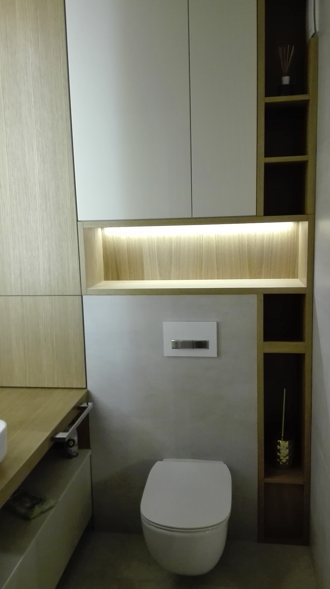 Nowoczesna łazienka z zabudową meblową w odcieniach bieli i drewna, podświetlana półka nad toaletą, minimalistyczny design.