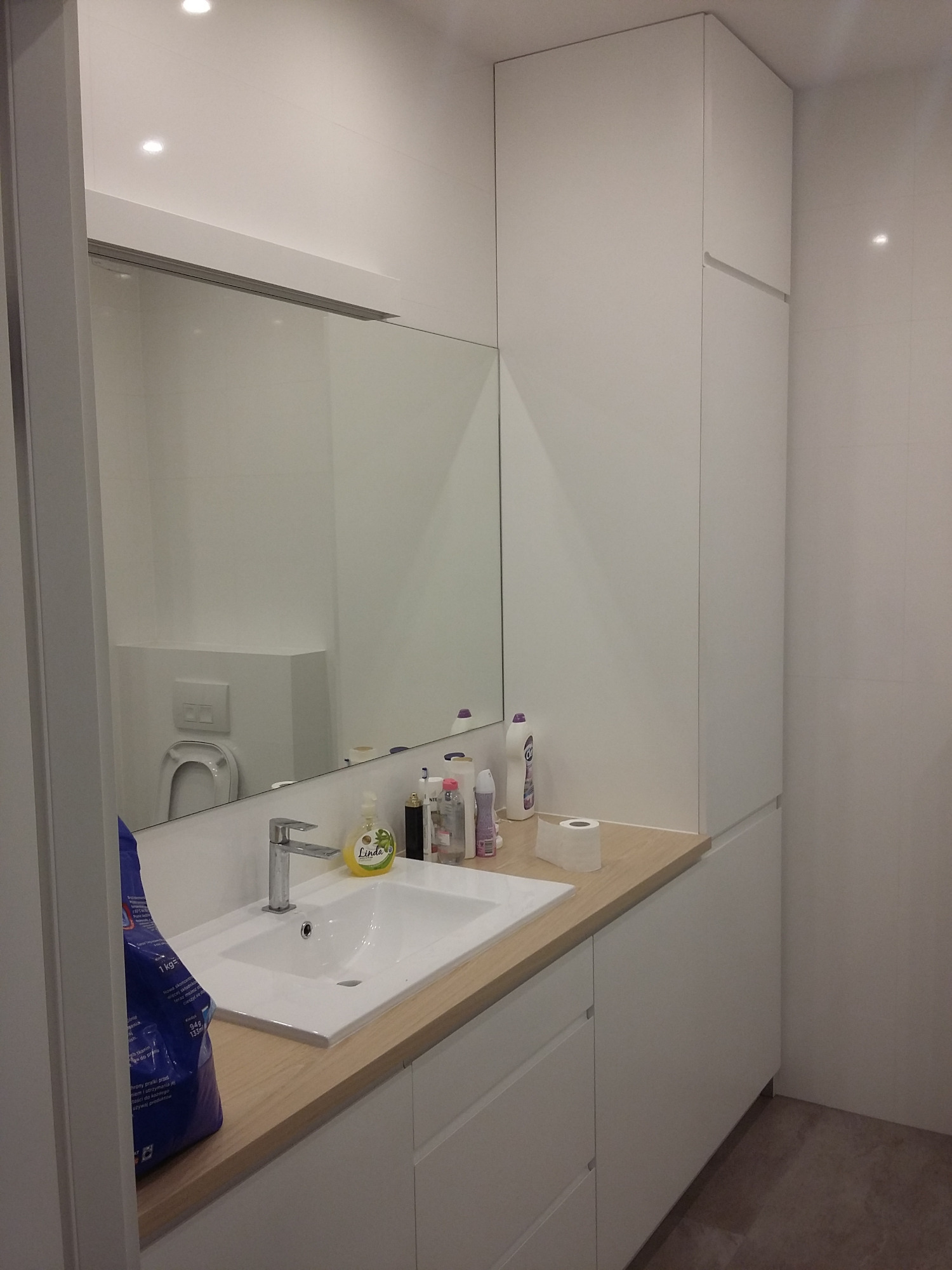 Nowoczesna łazienka z białą zabudową meblową, drewnianym blatem i dużym lustrem, w którym odbija się toaleta.