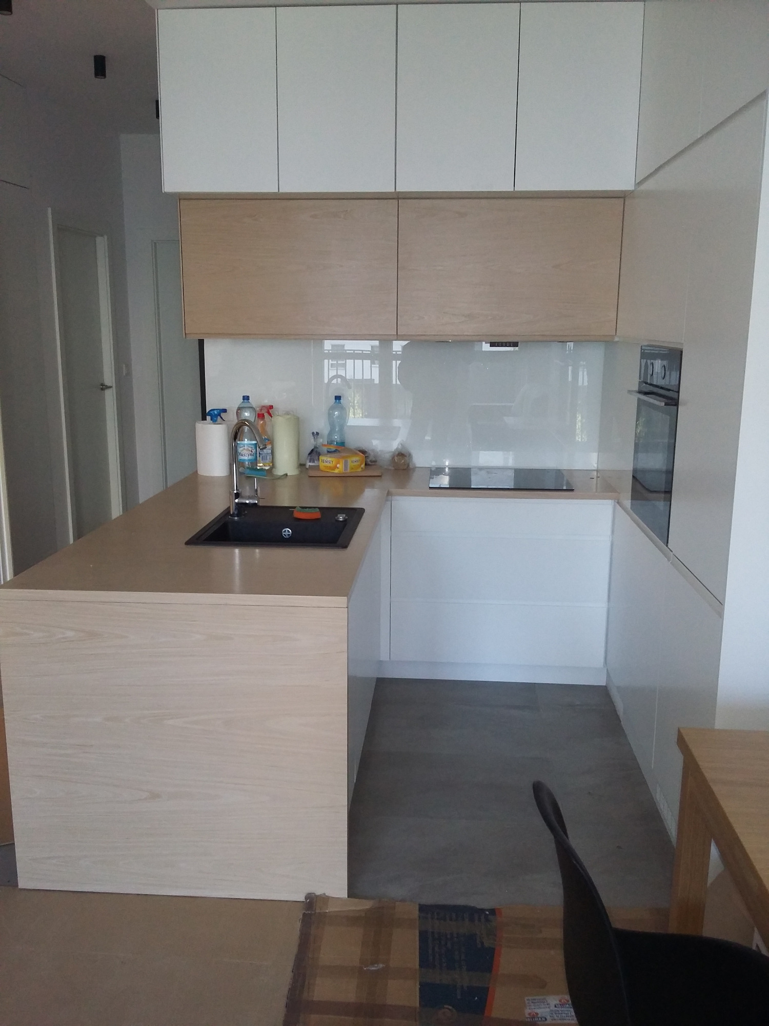 Nowoczesna kuchnia z jasnymi, drewnianymi frontami szafek i blatem, czarnym zlewem i białymi szafkami wiszącymi. Na blacie widoczne akcesoria kuchenne i środki czystości.