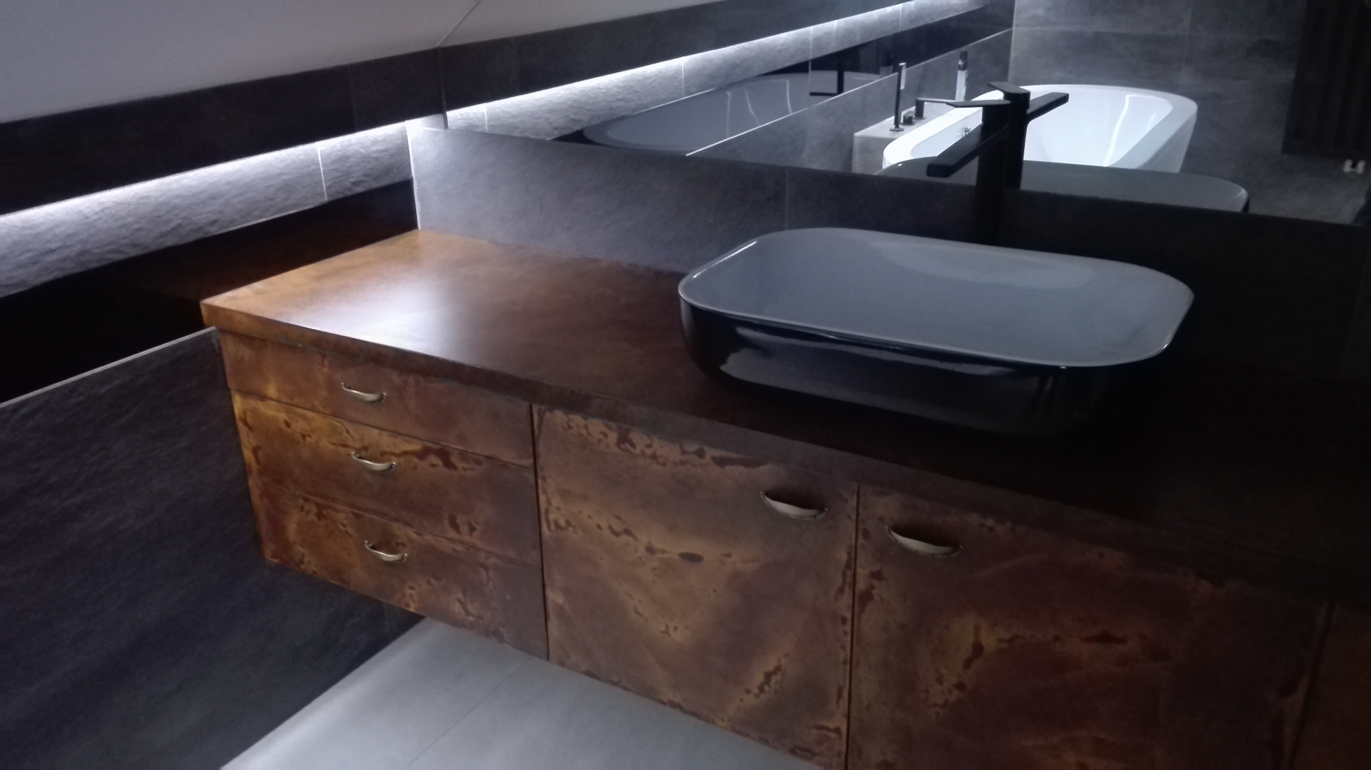 Nowoczesna łazienka z umywalką nablatową w kolorze czarnym i szafką pod umywalkę z brązowym, patynowanym wykończeniem, widoczne odbicie w lustrze z wanną i baterią.