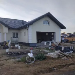 Ocieplony dom jednorodzinny z garażem w trakcie prac wykończeniowych elewacji, widoczne materiały budowlane i pracownicy w tle.