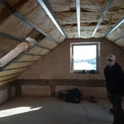 Poddasze w trakcie prac izolacyjnych: wełna mineralna między metalowymi profilami, widoczne okno z zimowym krajobrazem na zewnątrz, pracownik w maseczce i czarnym ubraniu.
