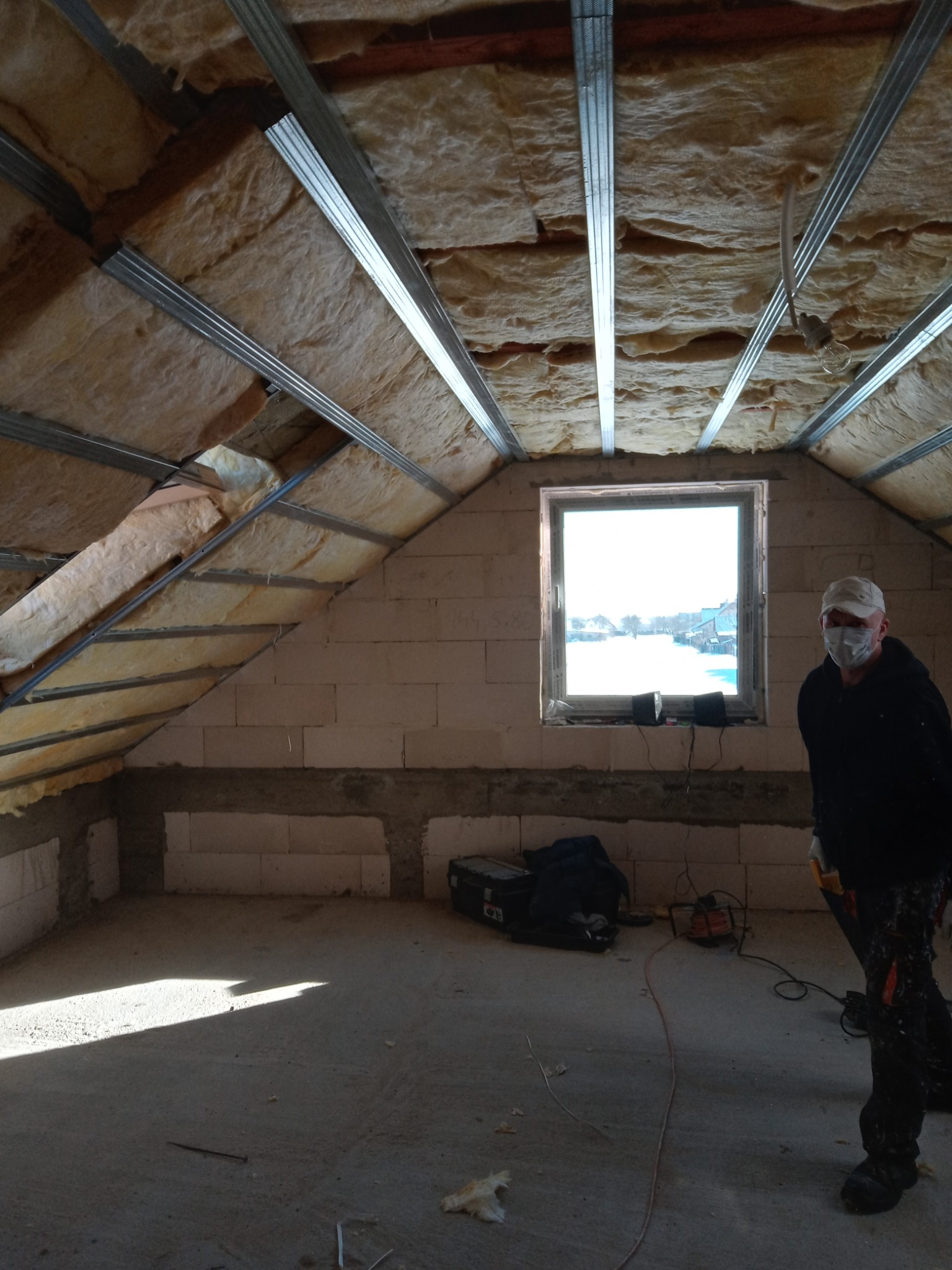 Poddasze w trakcie prac izolacyjnych: wełna mineralna między metalowymi profilami, widoczne okno z zimowym krajobrazem na zewnątrz, pracownik w maseczce i czarnym ubraniu.