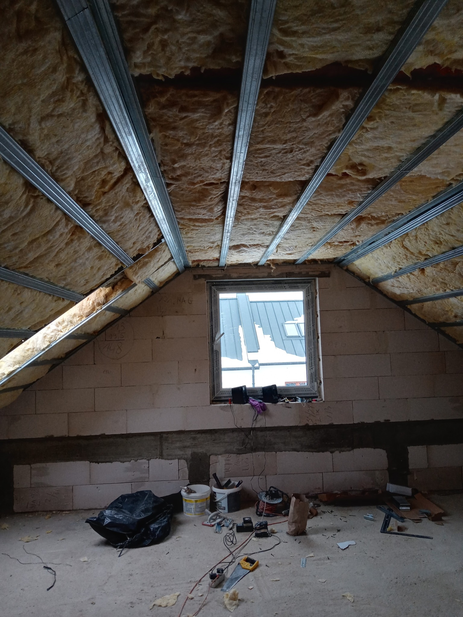 Poddasze w trakcie ocieplania: widoczna wełna mineralna między metalowymi profilami sufitowymi, okno z widokiem na dach i narzędzia na podłodze z betonu komórkowego.