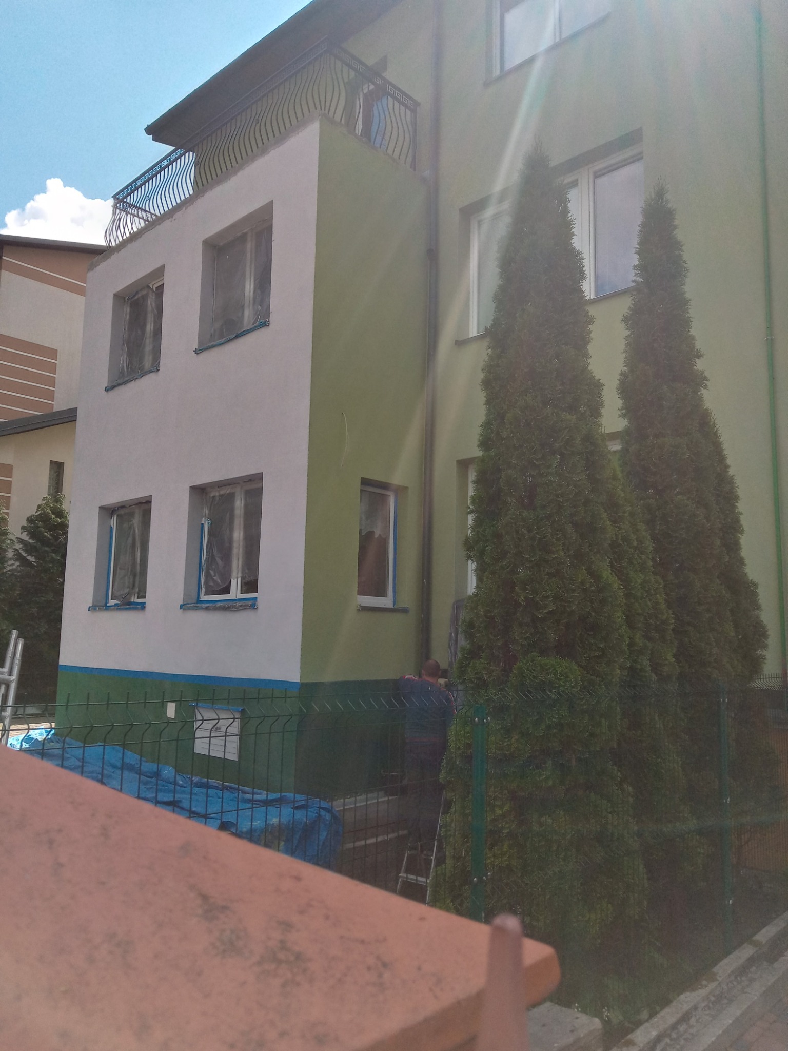 Dwukondygnacyjny budynek z ocieploną i pomalowaną elewacją w odcieniach szarości i zieleni, okna zabezpieczone folią malarską, widoczny pracownik na drabinie podczas prac wykończeniowych.