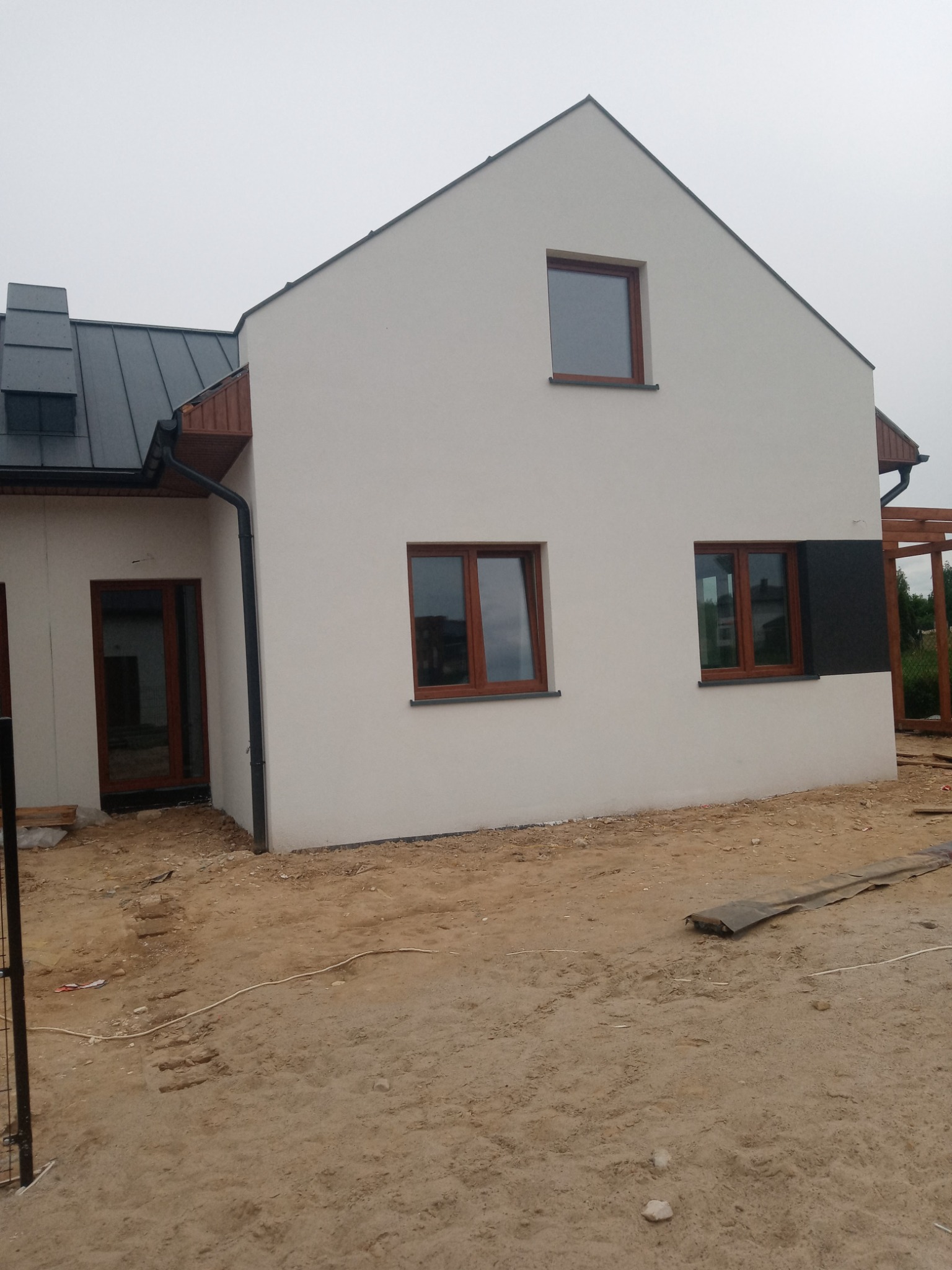Nowo wybudowany dom jednorodzinny z białą elewacją, brązowymi ramami okiennymi i szarą blachą na dachu, widoczny fragment czarnej izolacji elewacji z boku budynku i niezagospodarowany teren wokół.