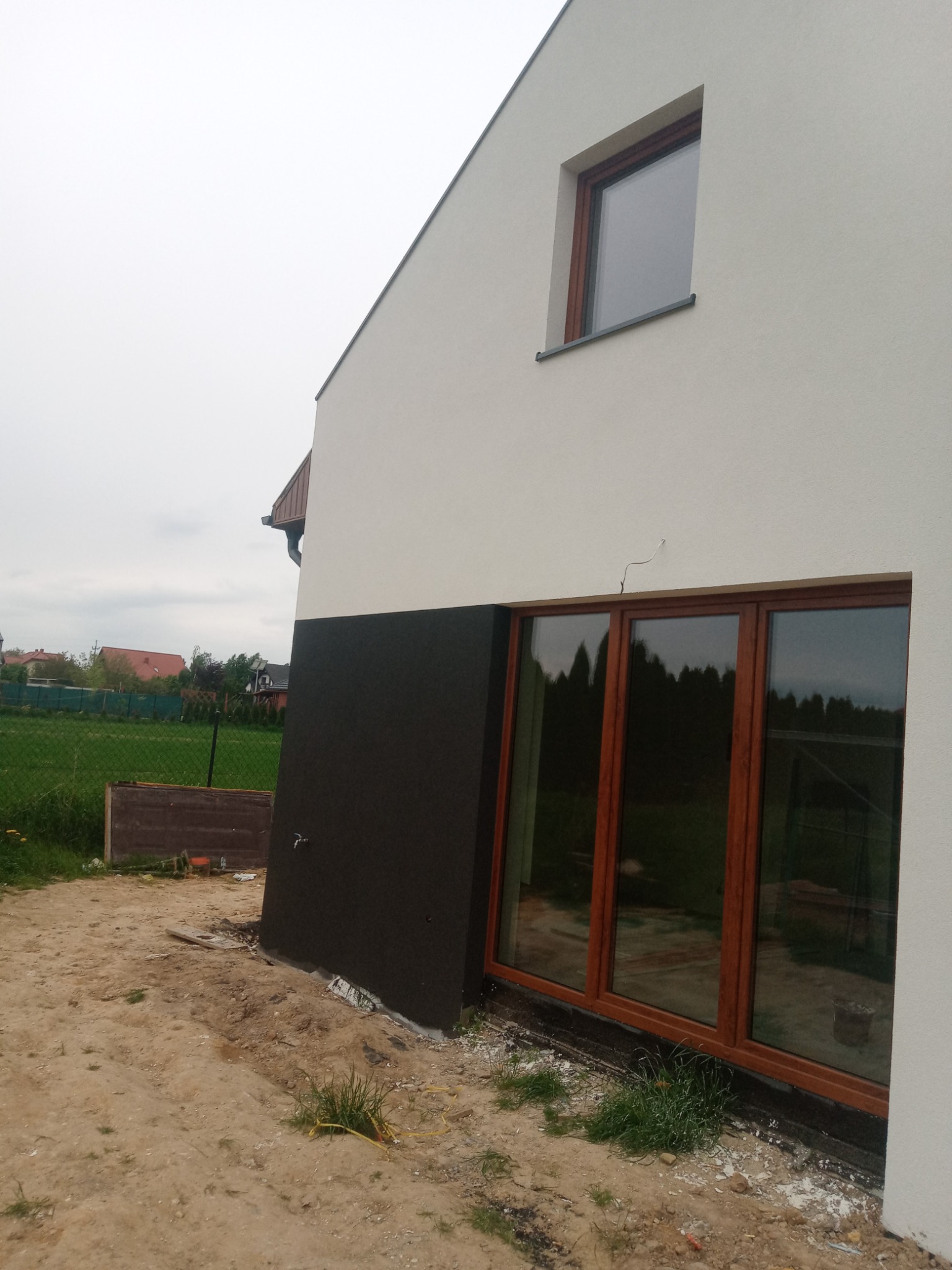 Fragment nowo wybudowanego domu z oknami w drewnianych ramach, z czarną, dolną częścią elewacji i jasną, górną częścią, otoczonego piaszczystym terenem.