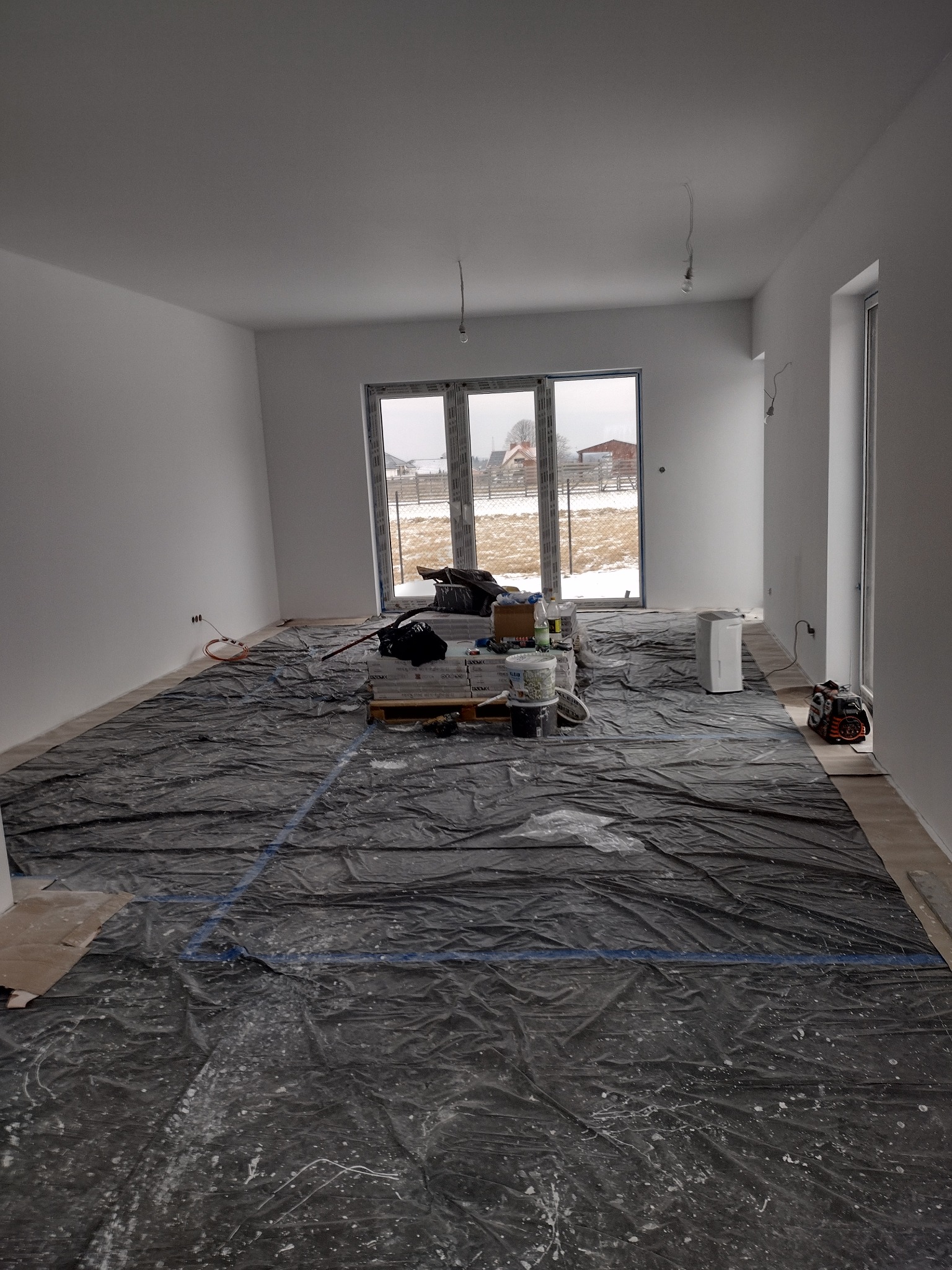 Wnętrze świeżo pomalowanego, jasnego pokoju zabezpieczone folią malarską, widoczne puszki z farbą, paleta paneli, osuszacz powietrza i narzędzia.