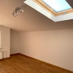 Puste, odnowione wnętrze z drewnianą podłogą, białymi ścianami, oknem dachowym i trzypunktową lampą sufitową, gotowe do aranżacji.