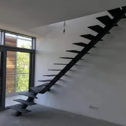 Nowoczesne, minimalistyczne schody stalowe bez poręczy, zamontowane przy ścianie w jasnym wnętrzu z dużym oknem.